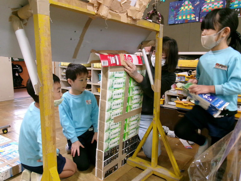 ケーキ屋さんの自動ドアを作りたいと始まりました。どうやったらドアが倒れないか？どうしたら人が入れるくらい開くようになるか？子どもたちは考えては試すを繰り返し、担任は子どもの思いを、うまくいかないかもしれないと思いながらも、まずはやらせて見守っています。