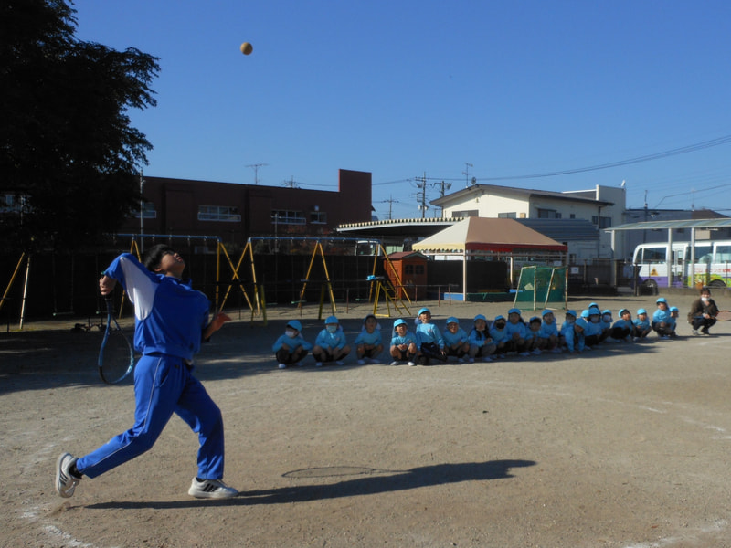 テニス部のお兄さん。職員が相手になってラリー。「これはサーブっていうんだよ」「どうやって打つの？」ボールが高く上がり子どもたちは心配していました。